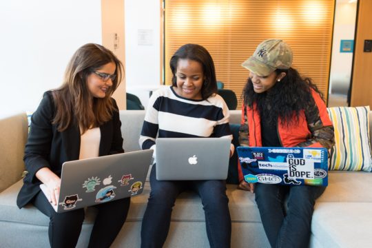 Three young professional women working together, sat on a sofa with Macbooks